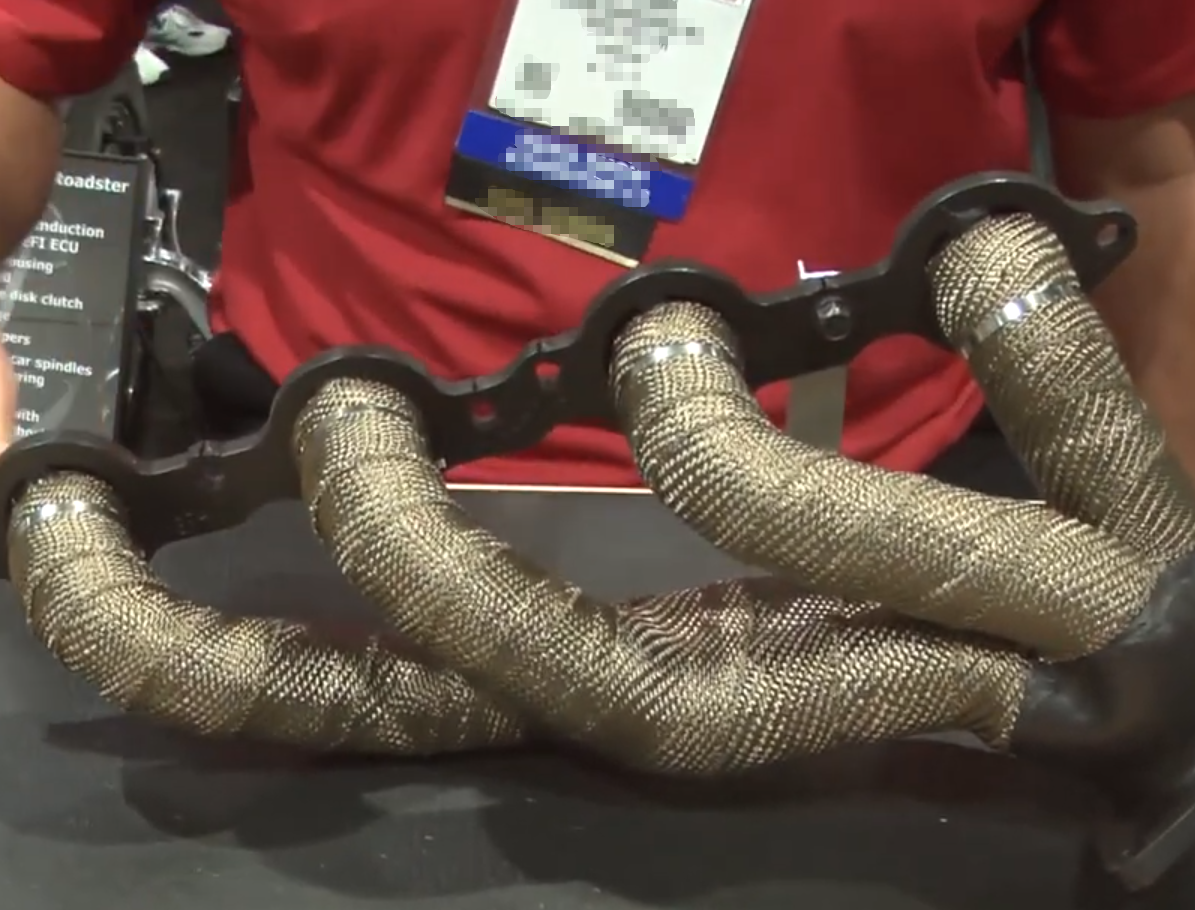 A close-up view of an exhaust manifold with four primary runners fully wrapped in gold-colored titanium (basalt) heat wrap, with stainless steel locking ties visible near the black mounting flange.